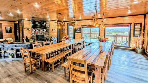 A rustic wooden interior with a long communal dining table, benches, and a fireplace.