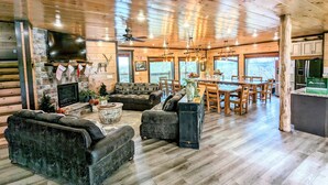 A spacious living room with a fireplace, a stone wall, and a large TV. There is a dining area with a wooden table and chairs.