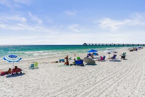 Ein Strand mit Menschen, die Sonnenbaden, Sonnenschirmen und einem Steg im Hintergrund.