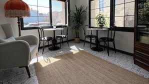 A room with a sofa, two tables with chairs, a potted plant, and large windows with a view of buildings outside.