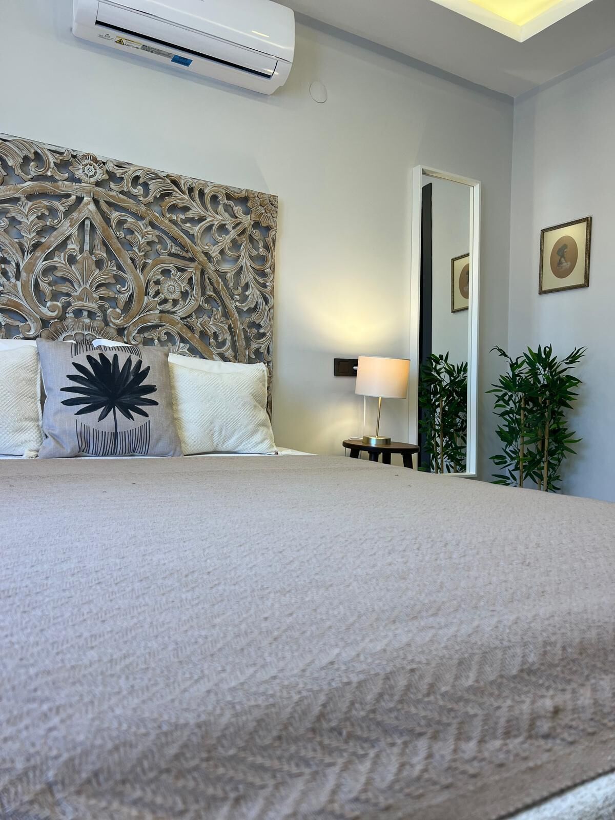 Una habitación de hotel con una cama grande, un equipo de aire acondicionado encima, un cabecero decorativo, una lámpara de noche y un espejo en la pared.