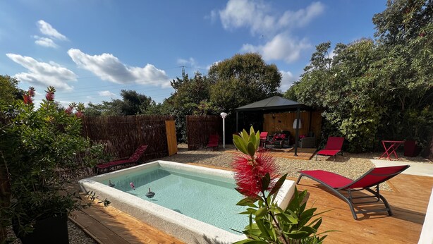 A backyard with a rectangular pool, surrounded by wooden decking, red lounge chairs, and a covered seating area. - La Maison de vos Vacances (Béziers)