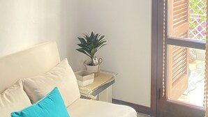 A living room with a sofa, a small table with a plant, a wall-mounted shelf with decorative items, and a large window with blinds. - Cozy holiday home in the heart of Versilia (Viareggio)