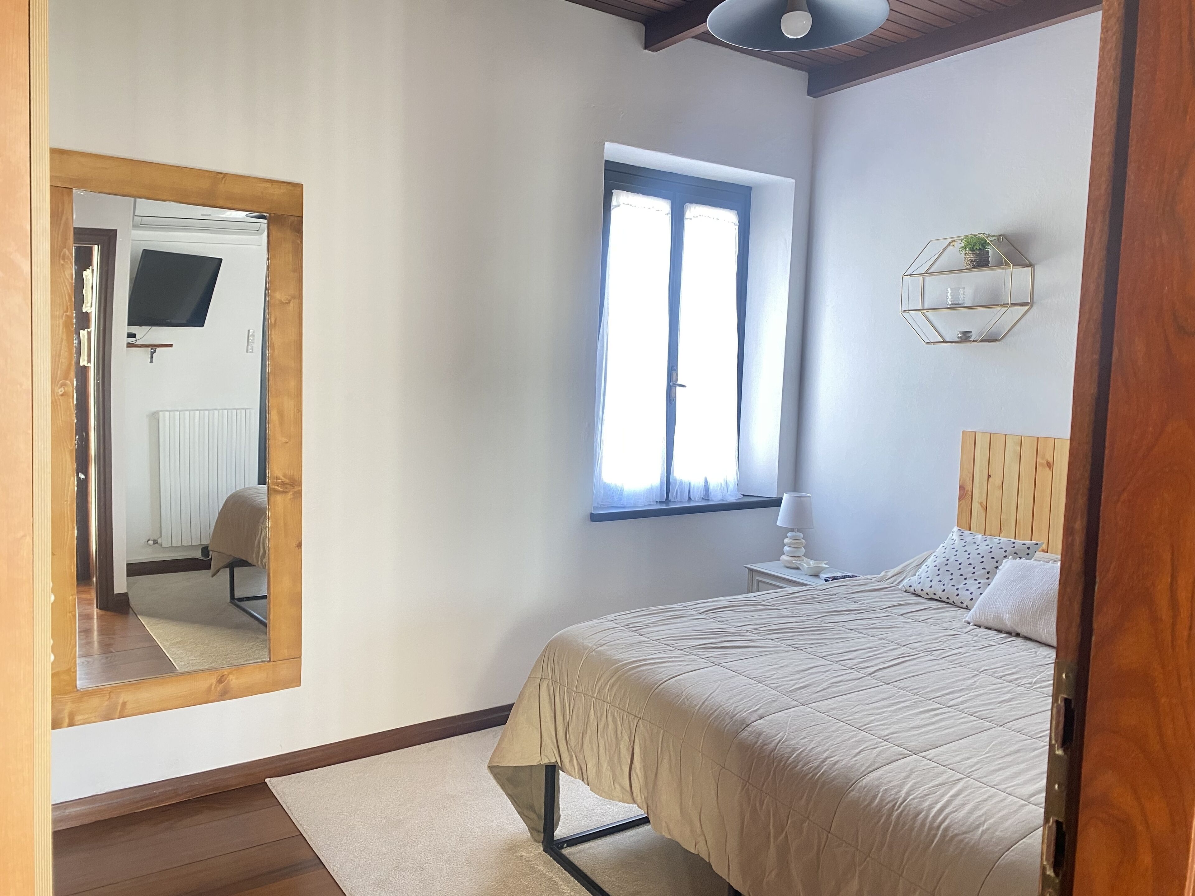 Un dormitorio con cama, ventana, espejo y mesita de noche con lámpara.