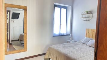 Un dormitorio con cama, ventana, espejo y mesita de noche con lámpara.