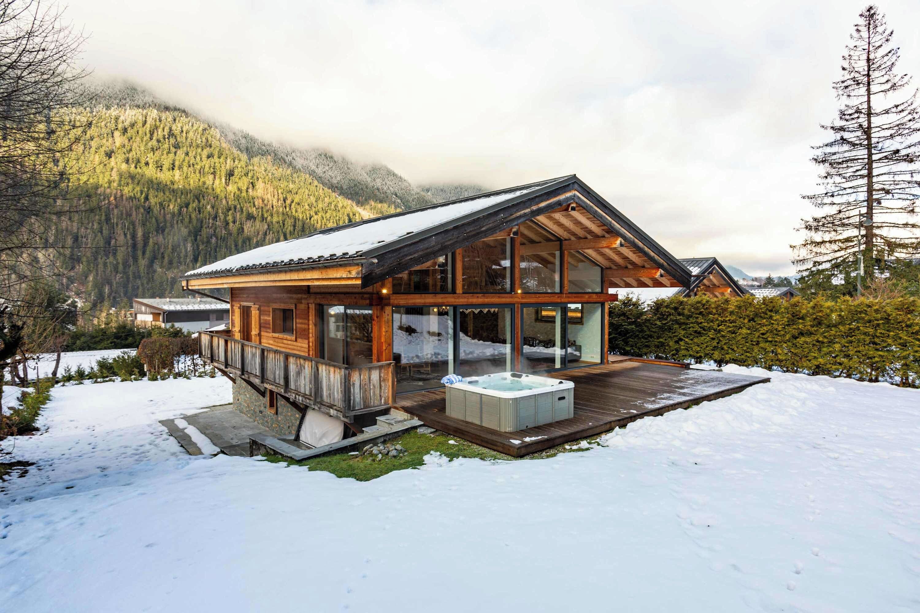 Uma cabana de madeira com uma extensão de vidro, uma banheira de hidromassagem e um deck rodeado por neve.