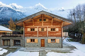 Een houten chalet met een stenen fundering, meerdere ramen en uitzicht op besneeuwde bergen.