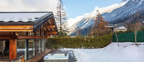 A modern cabin with a hot tub, wooden deck, and a view of snow-covered mountains.
