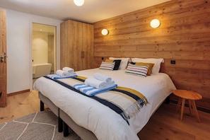 A bedroom with a wooden headboard, a bed with white bedding and striped towels, a wooden stool, and a wooden wardrobe.