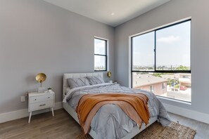A modern bedroom with a large bed, a nightstand, a lamp, and a view of the city through large windows. - Luxury Modern 4 Bed 3 Bath Home with Rooftop Deck #4 (Los Angeles)