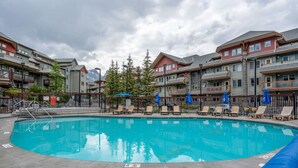 A swimming pool with lounge chairs and umbrellas, surrounded by multi-story buildings with balconies. - Spacious 3 Br | Heated Outdoor Pool + 3 Hot Tubs | Pool Table + Home Theatre (Canmore)