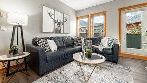A modern living room with a dark grey sofa, a round coffee table, and a large window with a mountain view. - Spacious 3 Br | Heated Outdoor Pool + 3 Hot Tubs | Pool Table + Home Theatre (Canmore)