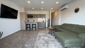 A modern living room with a green sofa, a flat-screen TV mounted on the wall, and a kitchen area with wooden cabinets and a stainless steel refrigerator.