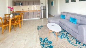 Ein modernes Wohnzimmer mit grauem Sofa, Esstisch aus Holz und Stühlen. Im Hintergrund ist eine Küche mit grauen Schränken und Edelstahlgeräten zu sehen.