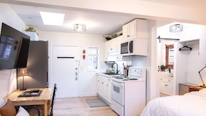 Cocina compacta con gabinetes blancos, un microondas y una estufa. Hay una zona de comedor con una mesa y sillas, y una cama con colcha blanca.