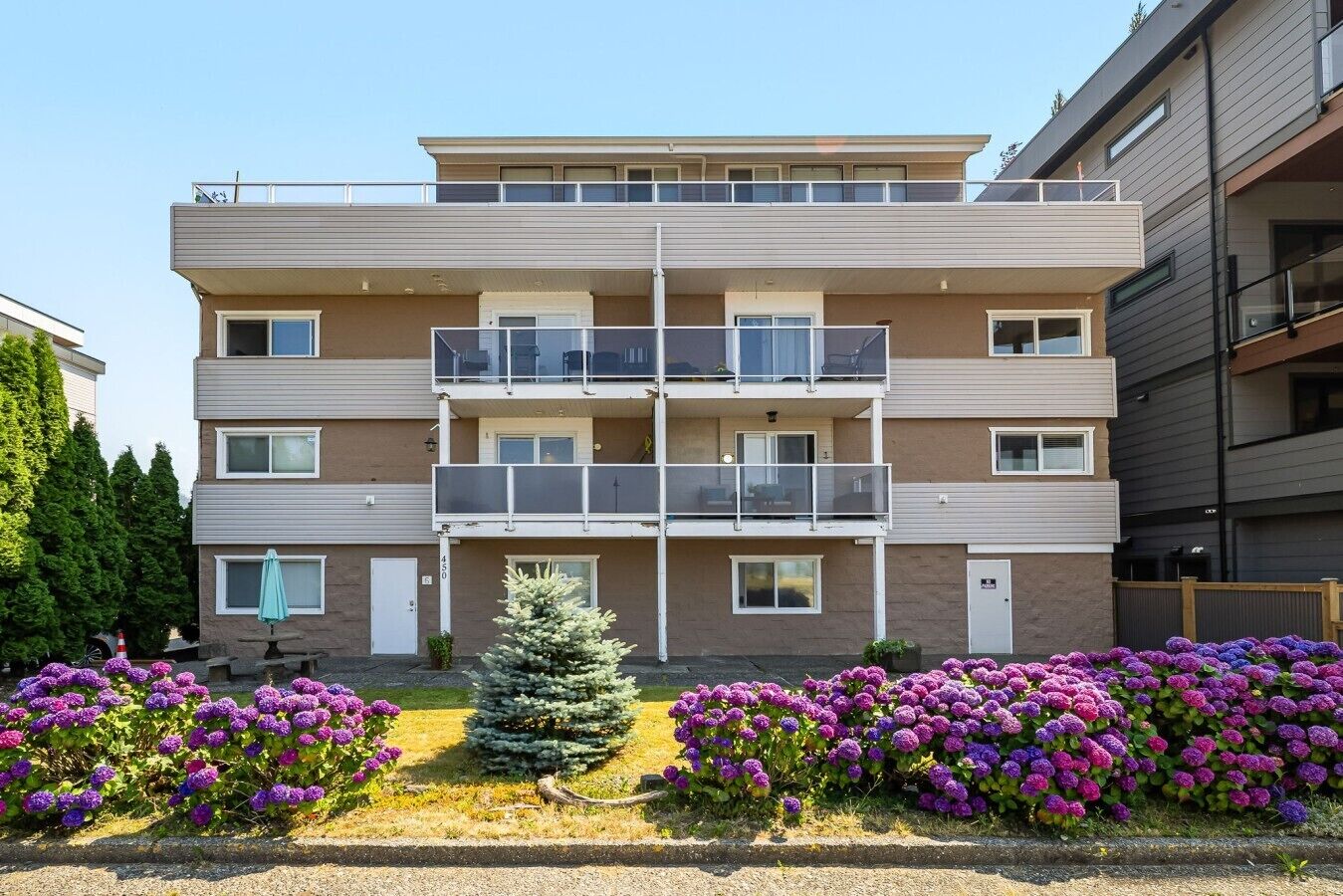 Un edificio residencial de varios pisos con balcones, un pequeño jardín con flores y cielo despejado.