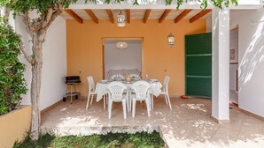 An outdoor patio with a table set for dining, surrounded by white chairs, under a pergola with climbing plants.