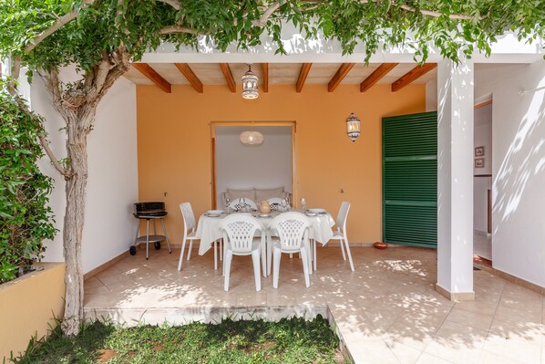 Um pátio externo com uma mesa posta para refeições, rodeado por cadeiras brancas, sob uma pérgola com plantas trepadeiras.