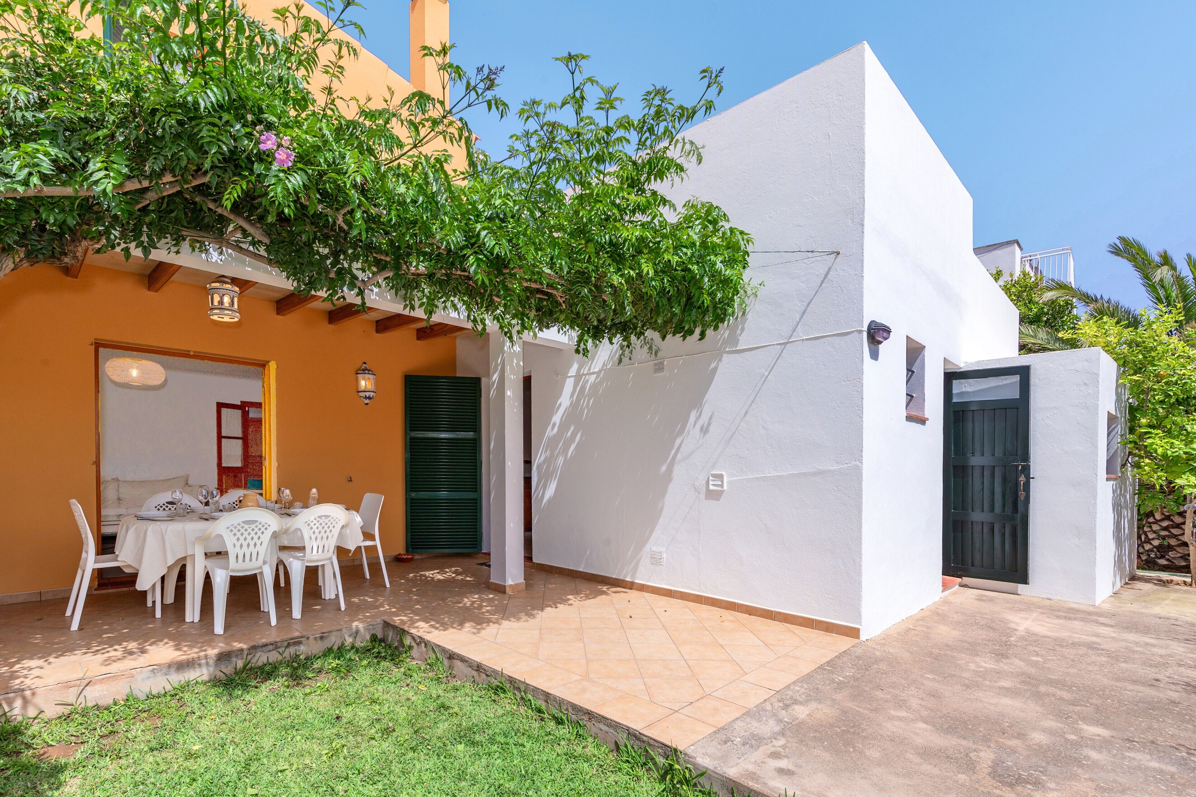 Un edificio de fachada blanca, con un patio que tiene una mesa y sillas, y un árbol de hojas verdes.
