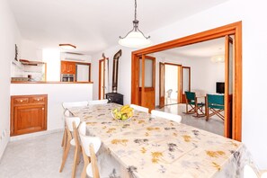 Uma área de refeições com uma mesa, cadeiras, uma cesta de frutas e uma cozinha ao fundo.