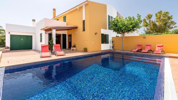 A swimming pool with a tiled deck, a covered patio area, and red lounge chairs.
