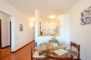 Un área de comedor con una mesa de vidrio, sillas de madera, un florero con una planta y una cocina al fondo.