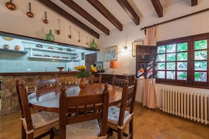 A dining area with a round wooden table and chairs, a kitchen with shelves of dishes and a radiator. - Villa 'Cuixach' with Private Pool, Wi-Fi and Air Conditioning (Pollensa)