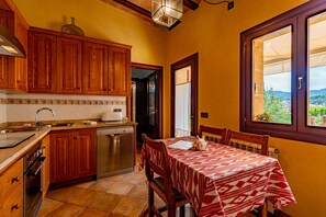 Une cuisine avec des meubles en bois, une table à manger recouverte d’une nappe à motifs et une vue par la fenêtre.