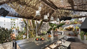 Un patio exterior con una mesa de madera, lámparas colgantes y varias macetas con plantas.