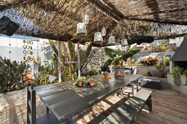 Un patio exterior con una mesa de madera, lámparas colgantes y varias macetas con plantas.