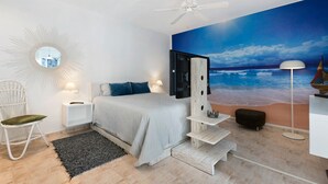 Un dormitorio con cama, una silla blanca, una mesita de noche, una lámpara, un ventilador de techo y un mural marino.