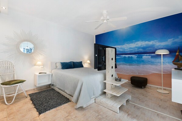 Quarto com cama, cadeira branca, mesa de cabeceira, abajur, ventoinha no teto e um mural na parede com tema oceânico.