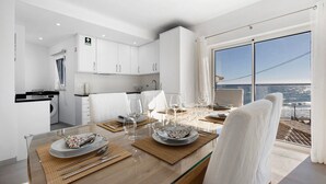 Un comedor moderno con vistas al mar, una cocina con armarios blancos y una mesa de comedor servida con platos y vasos.