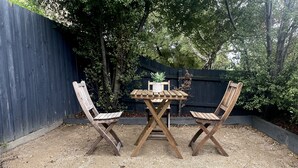 Un tavolo di legno e due sedie in un'area giardino con terreno sterrato, circondati da una recinzione in legno e alberi.