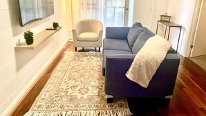 Un soggiorno con un divano grigio, una poltrona bianca, una televisione a schermo piatto montata a parete, un tappeto decorato e una piccola pianta su uno scaffale.