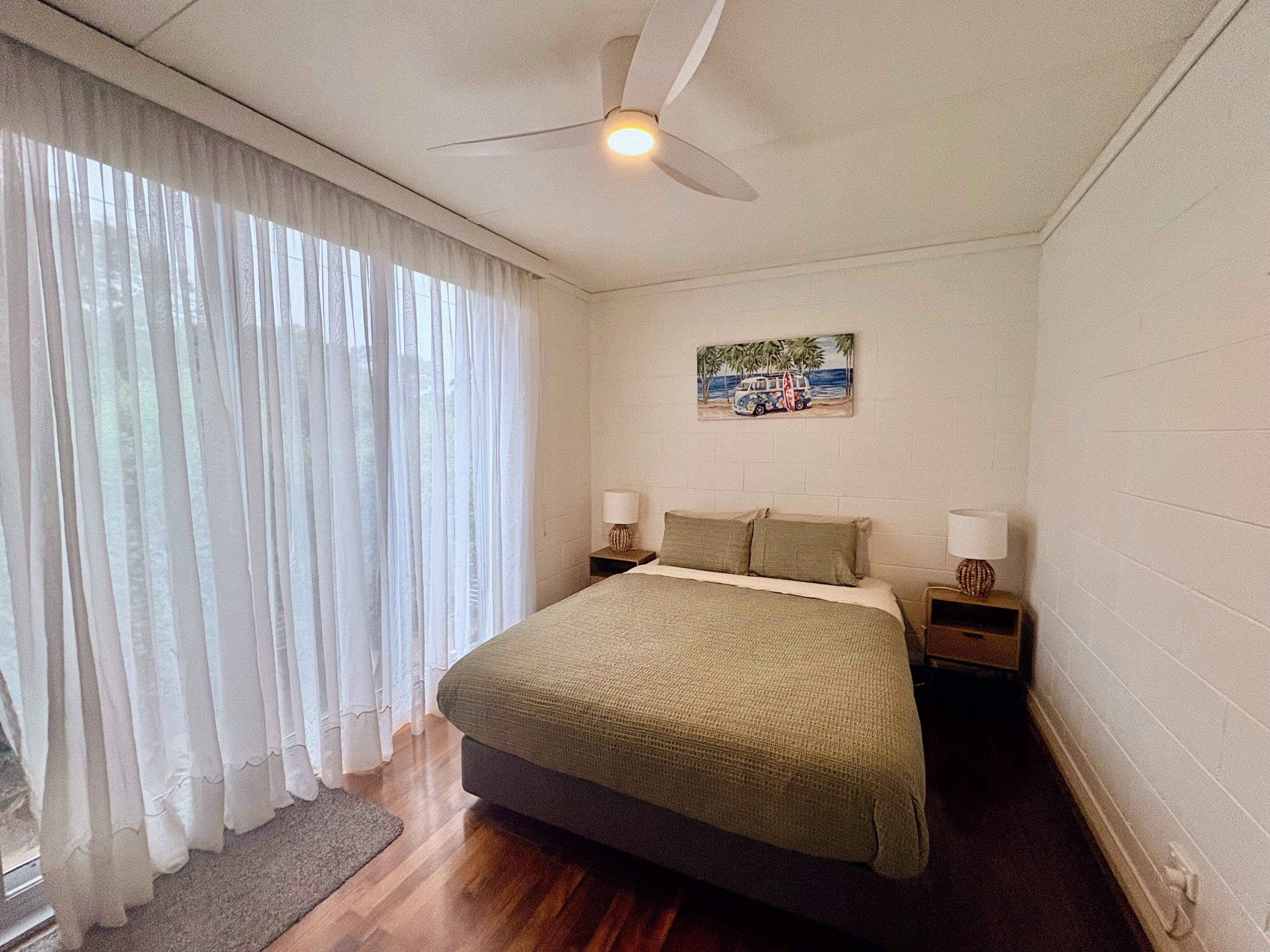 A bedroom with a bed, two bedside tables, a ceiling fan, and a window with sheer curtains.
