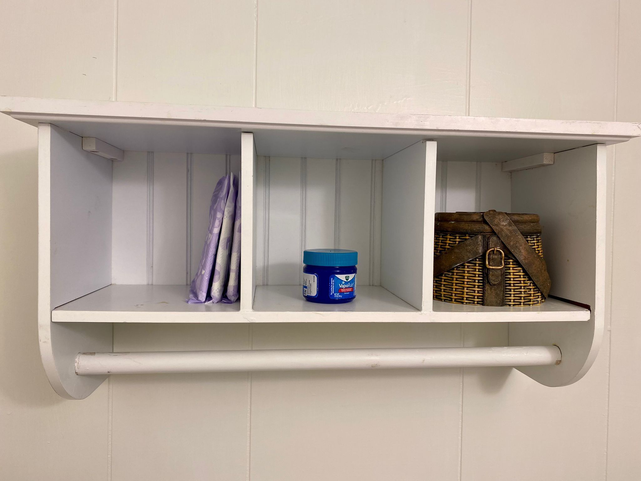 A white shelf with three compartments; one holds a blue container, another a purple cloth, and the third a wicker bag.