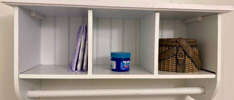 A white shelf with three compartments; one holds a blue container, another a purple cloth, and the third a wicker bag.