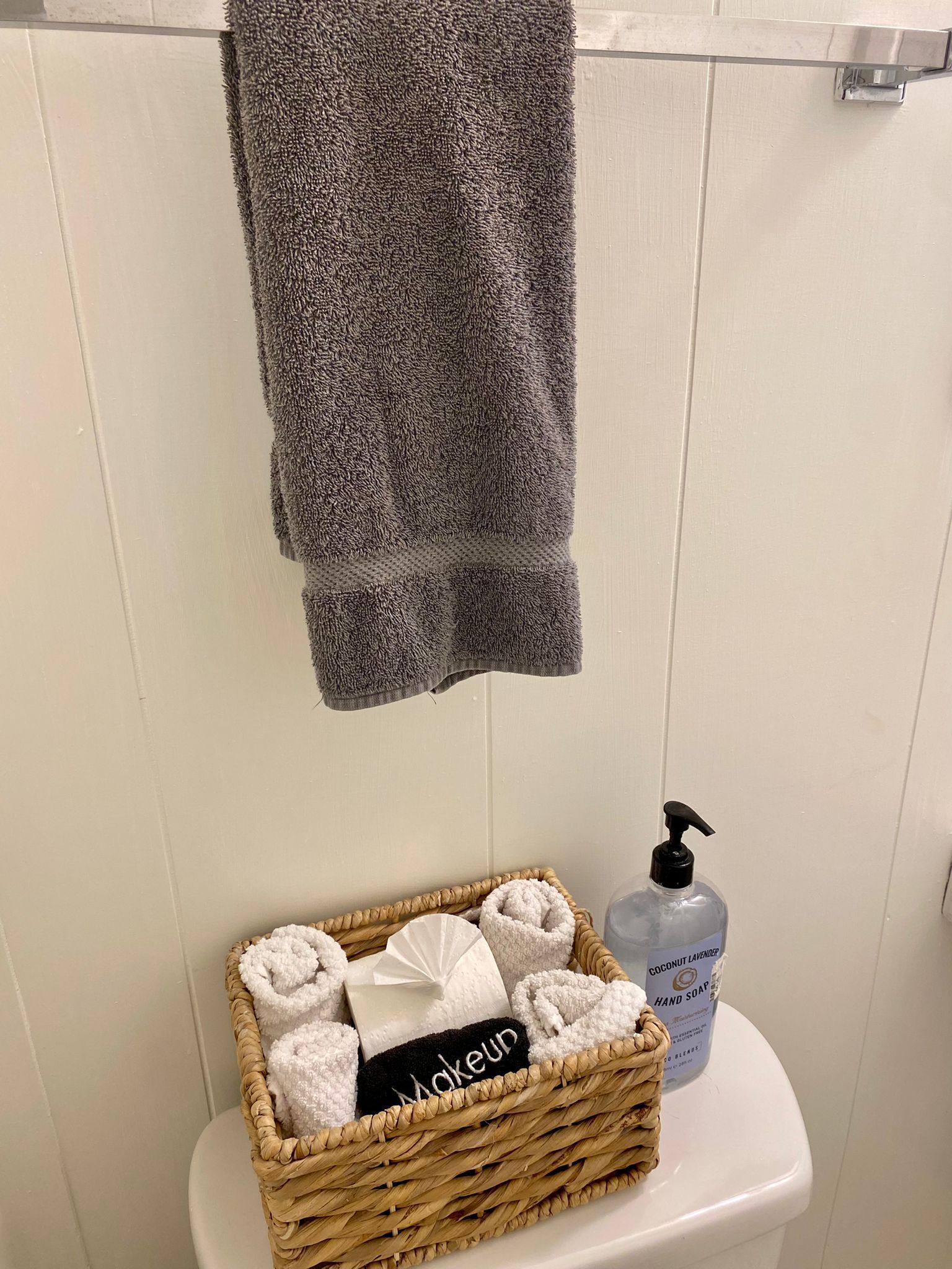 A bathroom with a white toilet, a wicker basket containing rolled towels, a bottle of hand soap, and a grey towel hanging on a metal towel rack.