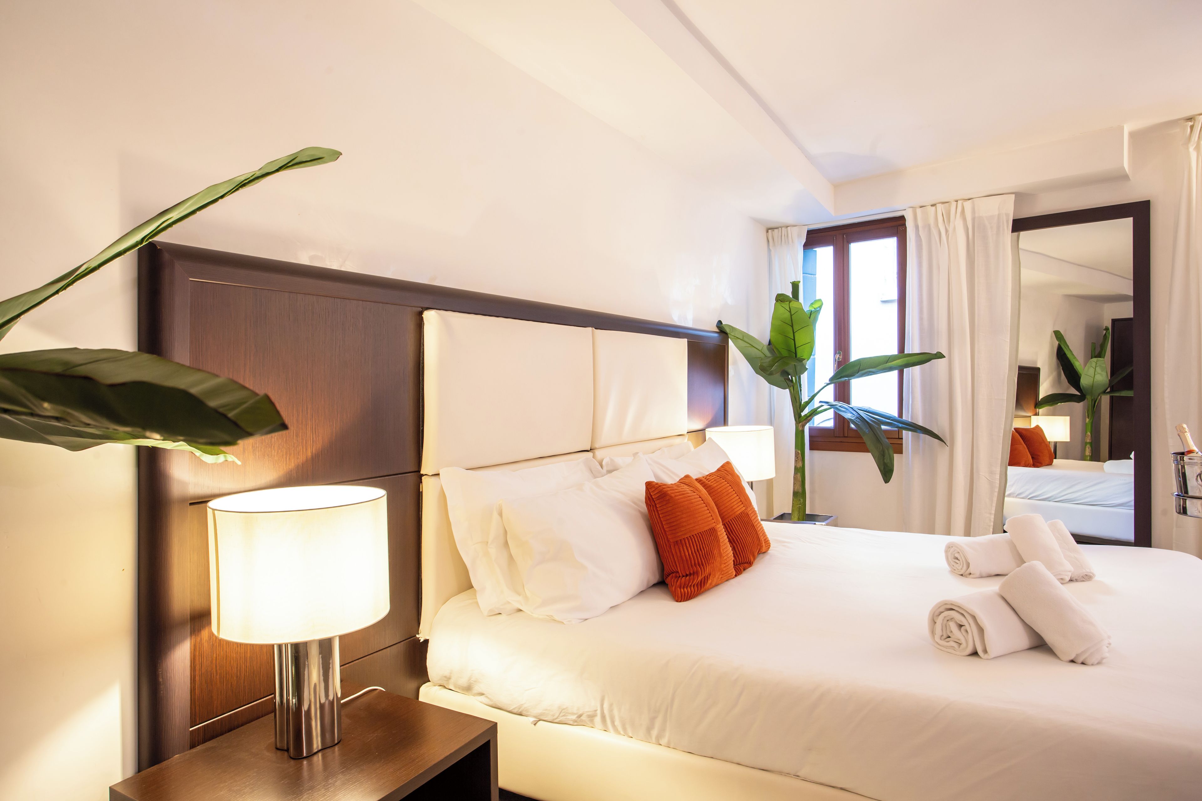 Habitación de hotel con cama, lámpara de noche, mesita de noche, espejo y ventana con cortinas.