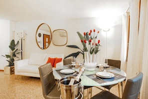 Un área de comedor con una mesa redonda preparada para dos personas, rodeada de sillas de cuero, un florero blanco con flores rojas y un cubo de champán plateado.