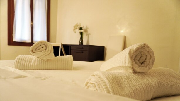 A neatly made bed with rolled towels and a vase of flowers on a nightstand.