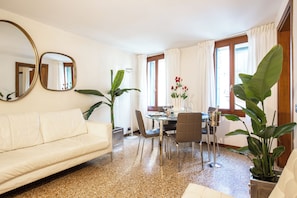 A modern living room with a white sofa, a dining table with chairs, and large windows with curtains.