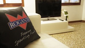 A living room with a white sofa, a black pillow with "ROGANTE Premium Sparkling Fruits" text, a flat-screen TV, and a small table with a remote control.