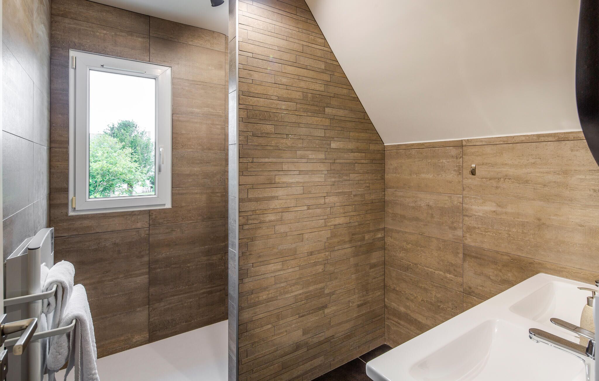 A modern bathroom with a sloped ceiling, a window with a view of greenery, a white bathtub, and a towel rack with towels.