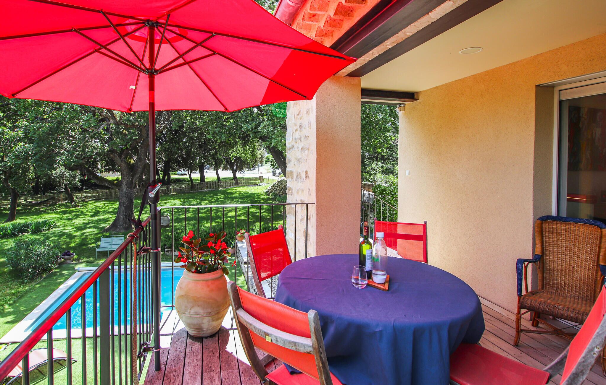 A balcony with a red umbrella, a round table with a blue tablecloth, wicker chairs, and a potted plant.