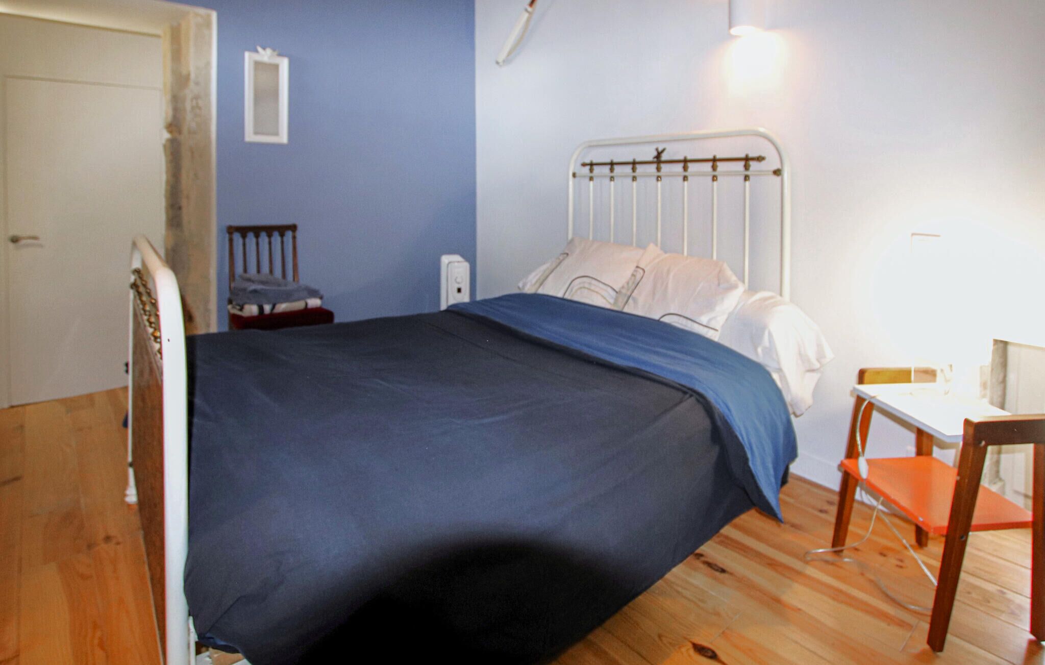 A bedroom with a metal bed frame, a blue bedspread, a wooden chair, and a small table.