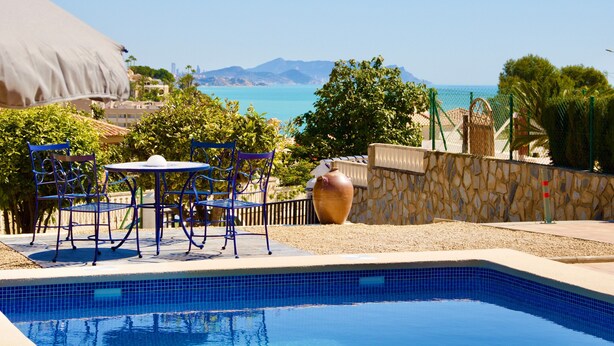 Ein Pool mit blauen Fliesen, ein Tisch mit Stühlen, eine große Vase und der Blick auf das Meer und die Berge.