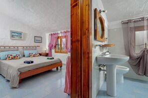A bedroom with a bed, a bedside table, a window with curtains, a bathroom with a sink and toilet, and a mirror. - Apartment in a villa surrounded by Gallura countryside, close to the sea (Olbia)