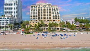 A beachfront hotel with a large building, palm trees, and a sandy beach with umbrellas.